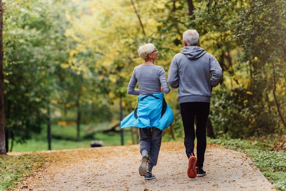 The back of an older couple walking/jogging through a park filled with trees
